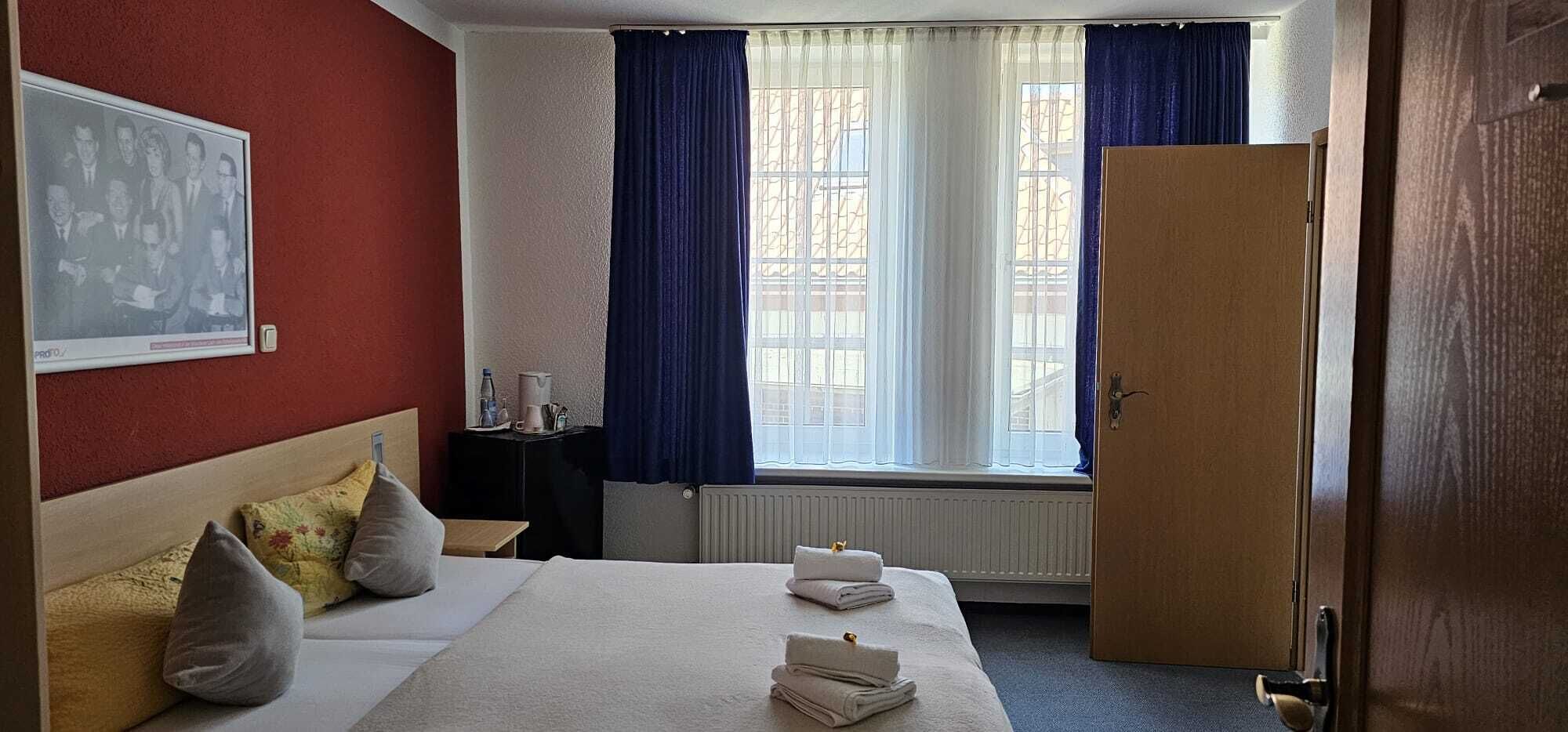 Habitación de hotel con cama, almohadas, mesita de noche, radiador, ventana con cortinas y puerta.
