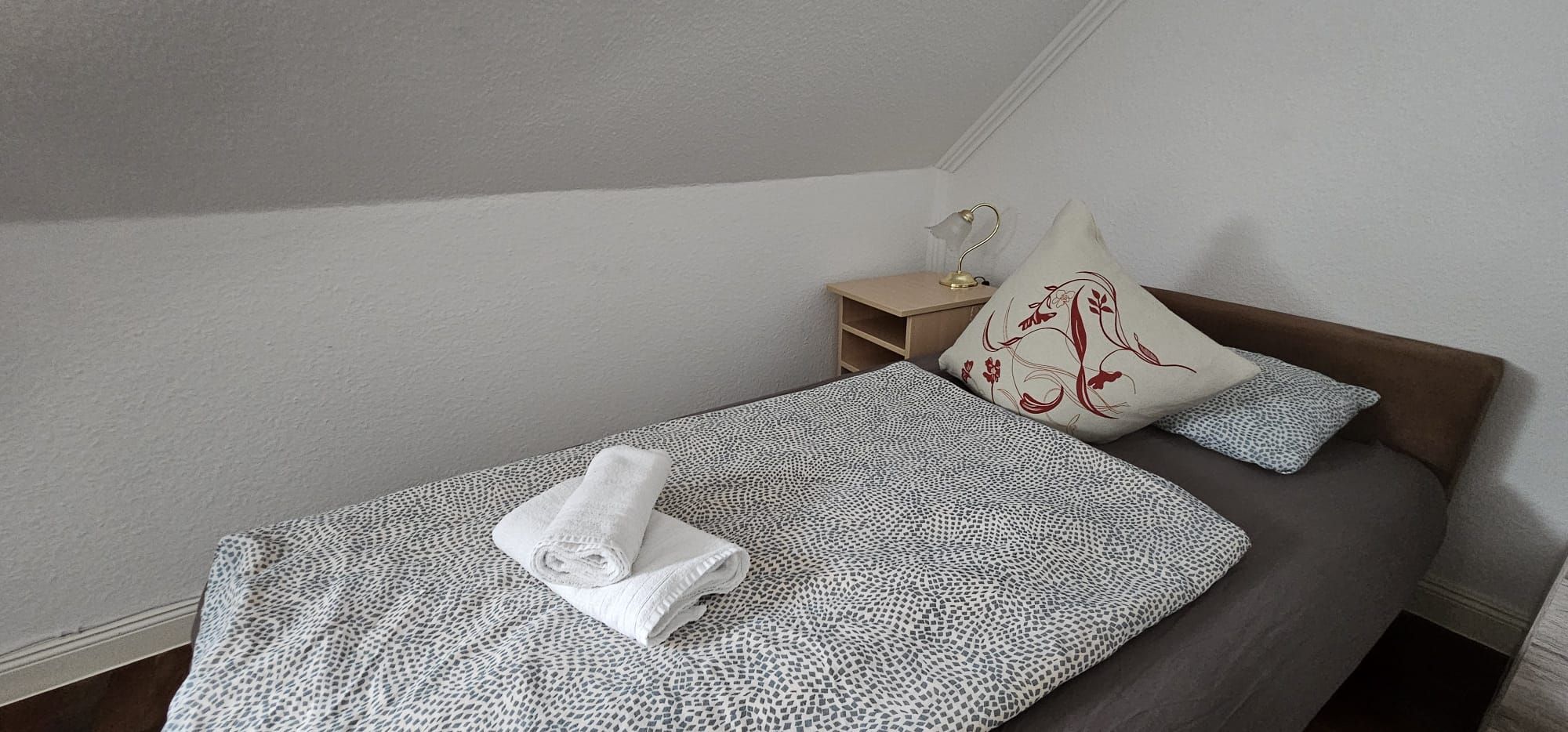 Ein Einzelbett mit einem gemusterten Bettüberwurf, ein zusammengerolltes Handtuch und zwei Kissen, eines mit einem Blumenmuster.