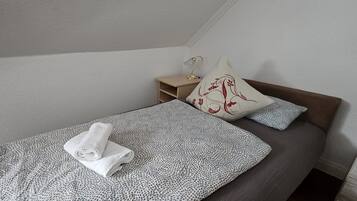 Ein Einzelbett mit einem gemusterten Bettüberwurf, ein zusammengerolltes Handtuch und zwei Kissen, eines mit einem Blumenmuster.