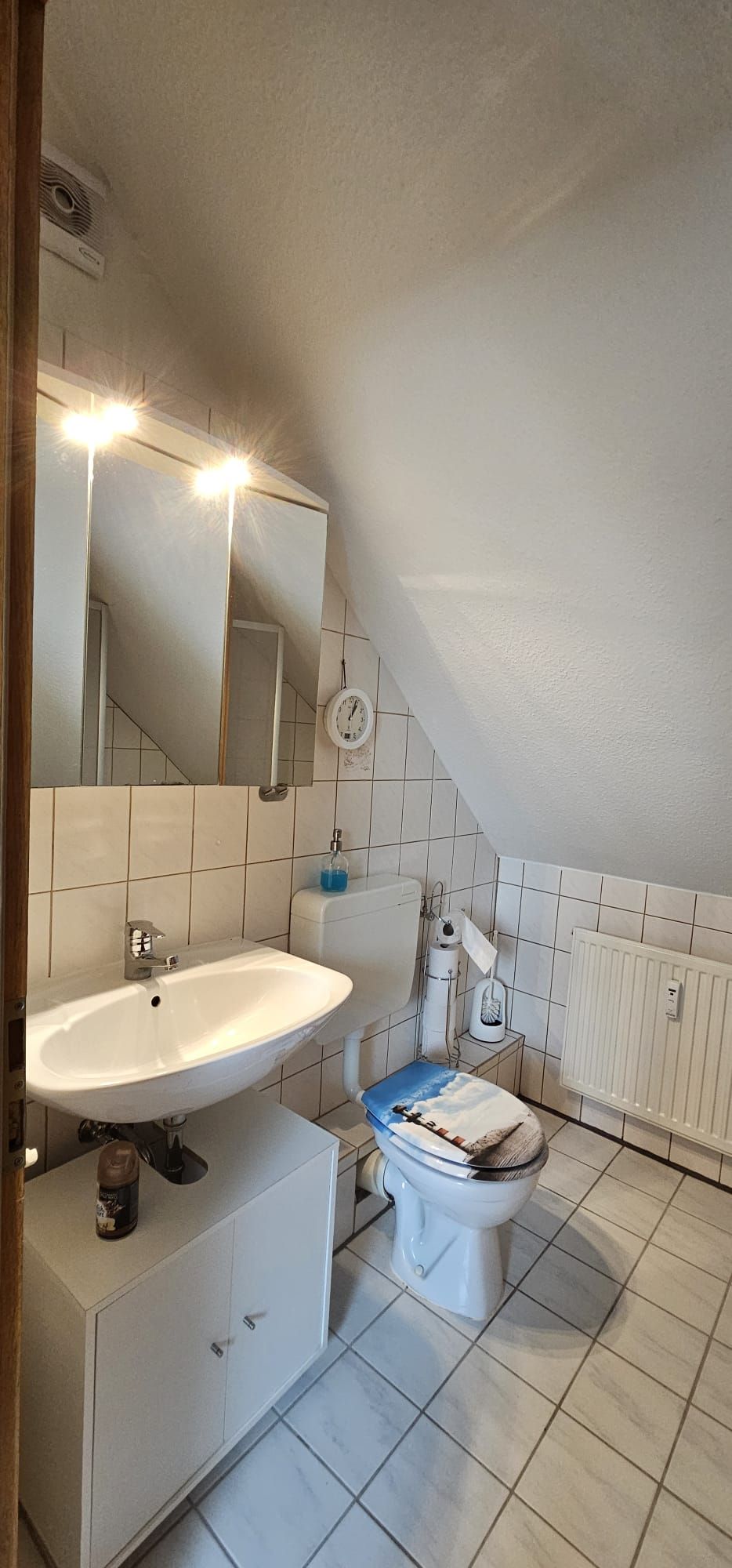 Un baño con un lavabo blanco, un inodoro con tapa azul, un espejo y un reloj en la pared.