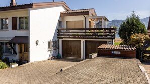 A two-story house with a garage, a balcony with plants, and a view of the mountains. - Luna Appartement/Fewo 2-4 Bettwohnung by Interhome (Gwatt (Thun))
