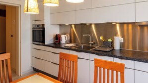 A modern kitchen with a stainless steel backsplash, a built-in oven, a coffee maker, and a sink. There are wooden chairs and a table in the dining area. - Hauswurz Appartement/Fewo Appartement/Fewo by Interhome (Gwatt (Thun))