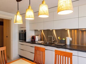 A modern kitchen with a stainless steel backsplash, a built-in oven, a coffee maker, and a sink. There are wooden chairs and a table in the dining area. - Hauswurz Appartement/Fewo Appartement/Fewo by Interhome (Gwatt (Thun))