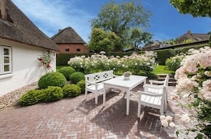 Eine Terrasse mit weißem Tisch und Stühlen, umgeben von üppiger Bepflanzung und blühenden Blumen.