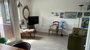 Uma sala de estar com uma mesa de madeira, uma cadeira verde, um sofá, uma televisão sobre um móvel e uma cadeira com almofada, além de obras de arte na parede.