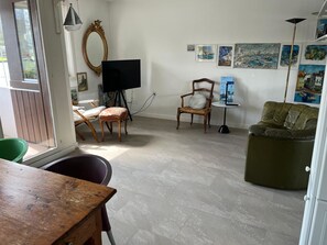 Ein Wohnzimmer mit Holztisch, einem grünen Sessel, einer Couch, einem Fernseher auf einem Standfuß, einem Sessel mit Kissen und Kunstwerken an der Wand.