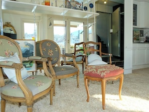 A room with antique-style furniture, including chairs and a table, a large window with a view, and a kitchen area.