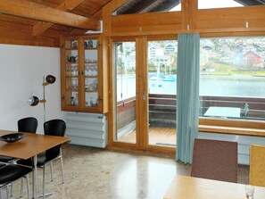 Ein Zimmer mit Holzboden, einem Esstisch mit Stühlen, einer Vitrine mit Glastüren und einem großen Fenster mit Blick auf ein Gewässer und Gebäude.