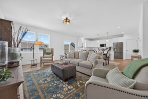 Ein modernes Wohnzimmer mit Sofa, Hocker und Couchtisch, ein Essbereich mit Stühlen und eine Küche mit Geräten.