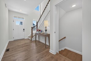 Ein Flur mit Holzboden, einer weißen Tür, einer Treppe mit hölzernem Geländer sowie einem Tisch mit Lampe und Blumen.