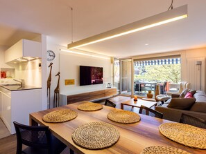 Eine moderne Küche mit Einbauofen, ein Essbereich mit Holztisch und gewebten Platzsets, eine Couch und ein an der Wand befestigter Fernseher.