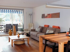 Ein modernes Wohnzimmer mit einer Couch, einem Esstisch und einem Sessel. Ein Fenster bietet Blick ins Freie.