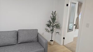 En grå soffa i ett rum med vita väggar, en krukväxt och en dörr som leder till ett annat rum.