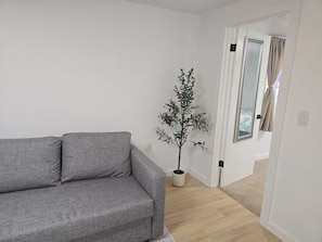 A grey sofa in a room with white walls, a potted plant, and a door leading to another room. - Studio Near Airport & City - Cozy Green Retreat! (Seattle)