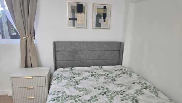 Ett sovrum med en grå sänggavel, en säng med ett sängöverkast med ett lövmönster, ett sängbord med lådor och två abstrakta konstverk på väggen.