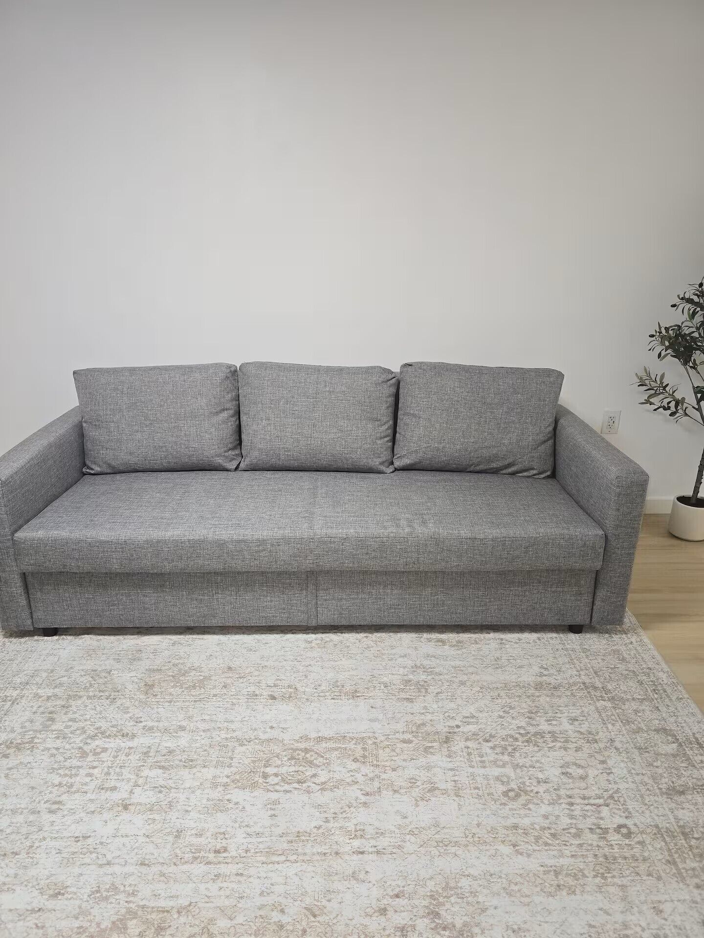A grey sofa with three cushions against a plain wall, a white rug, and a potted plant to the right.