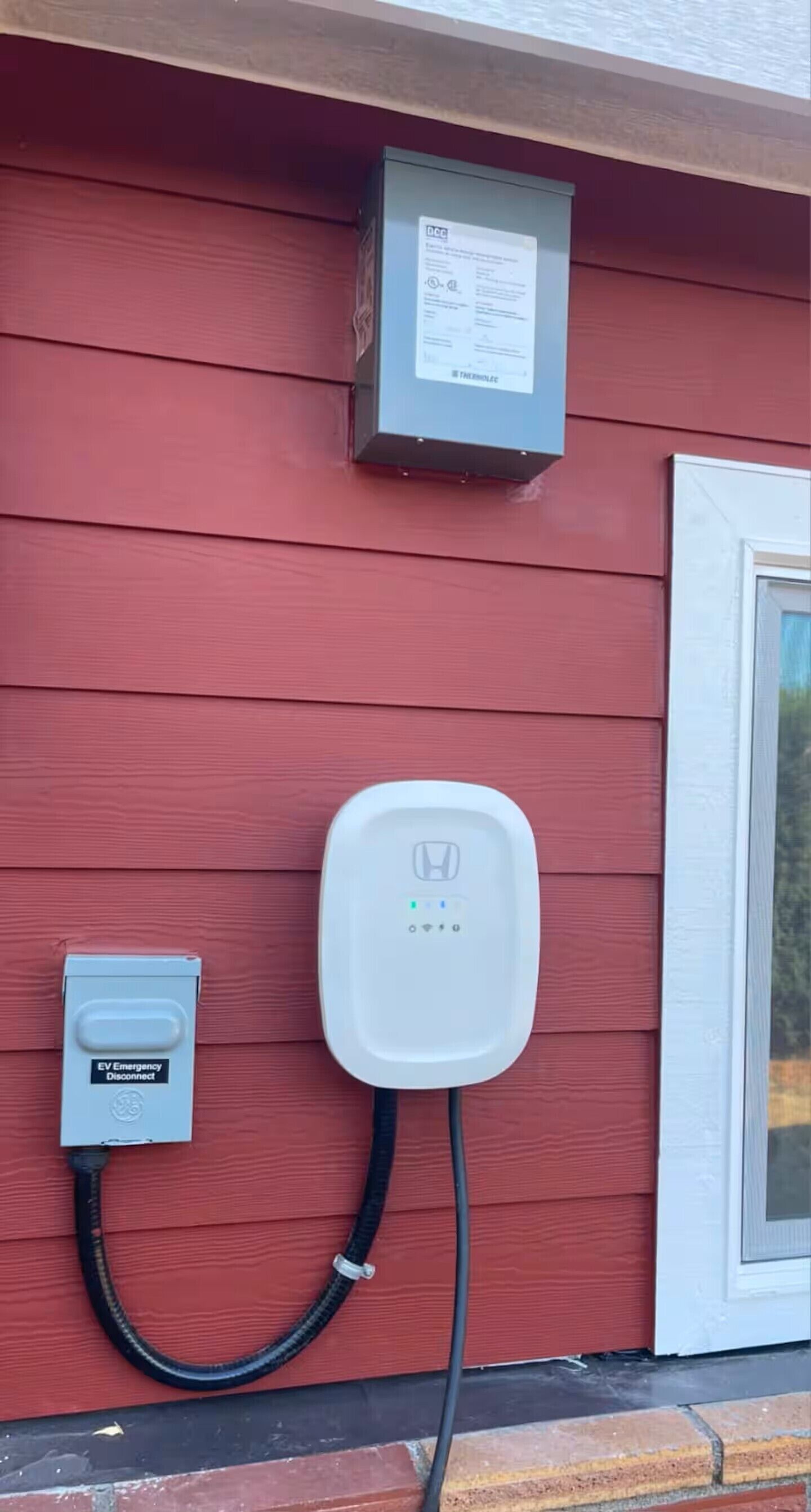 A red building with a white door, a white electrical box, and a white electrical outlet with a black cable connected.