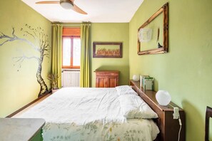 Un dormitorio con cama, mesita de noche de madera, ventilador de techo, radiador, ventana con cortinas y un cuadro en la pared.