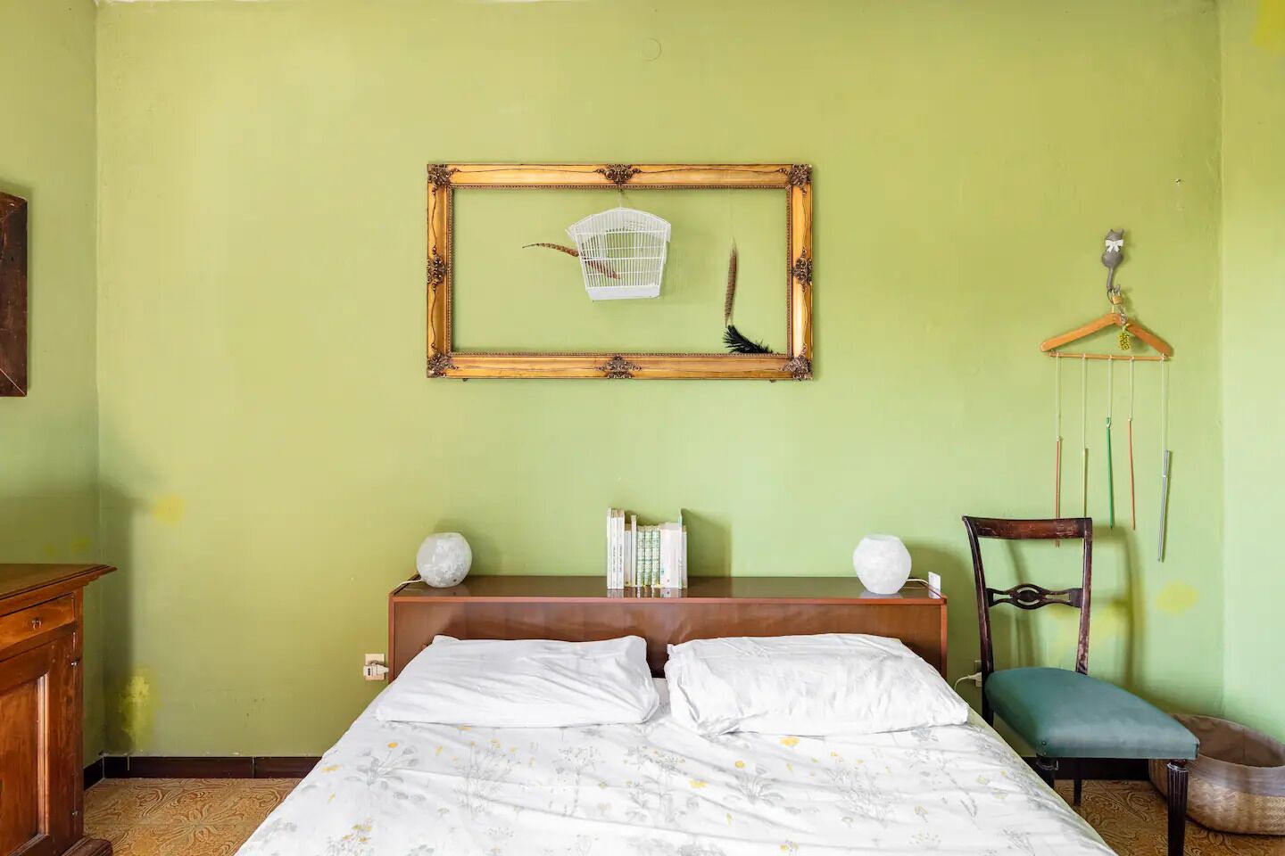 A room with a bed, a wooden dresser, a chair, a hanging birdcage, and a framed mirror on the wall.