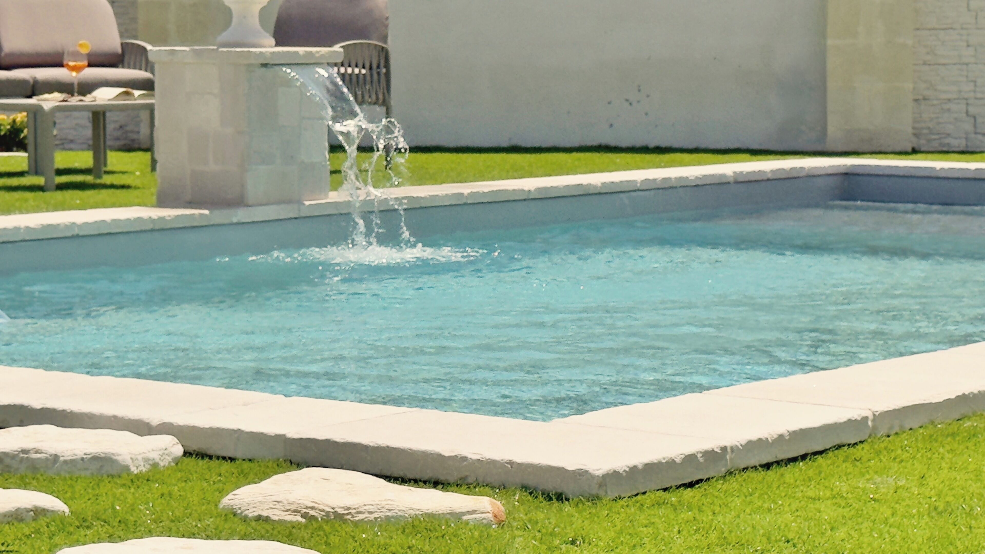 A rectangular swimming pool with clear blue water, surrounded by a stone border and a grassy area.