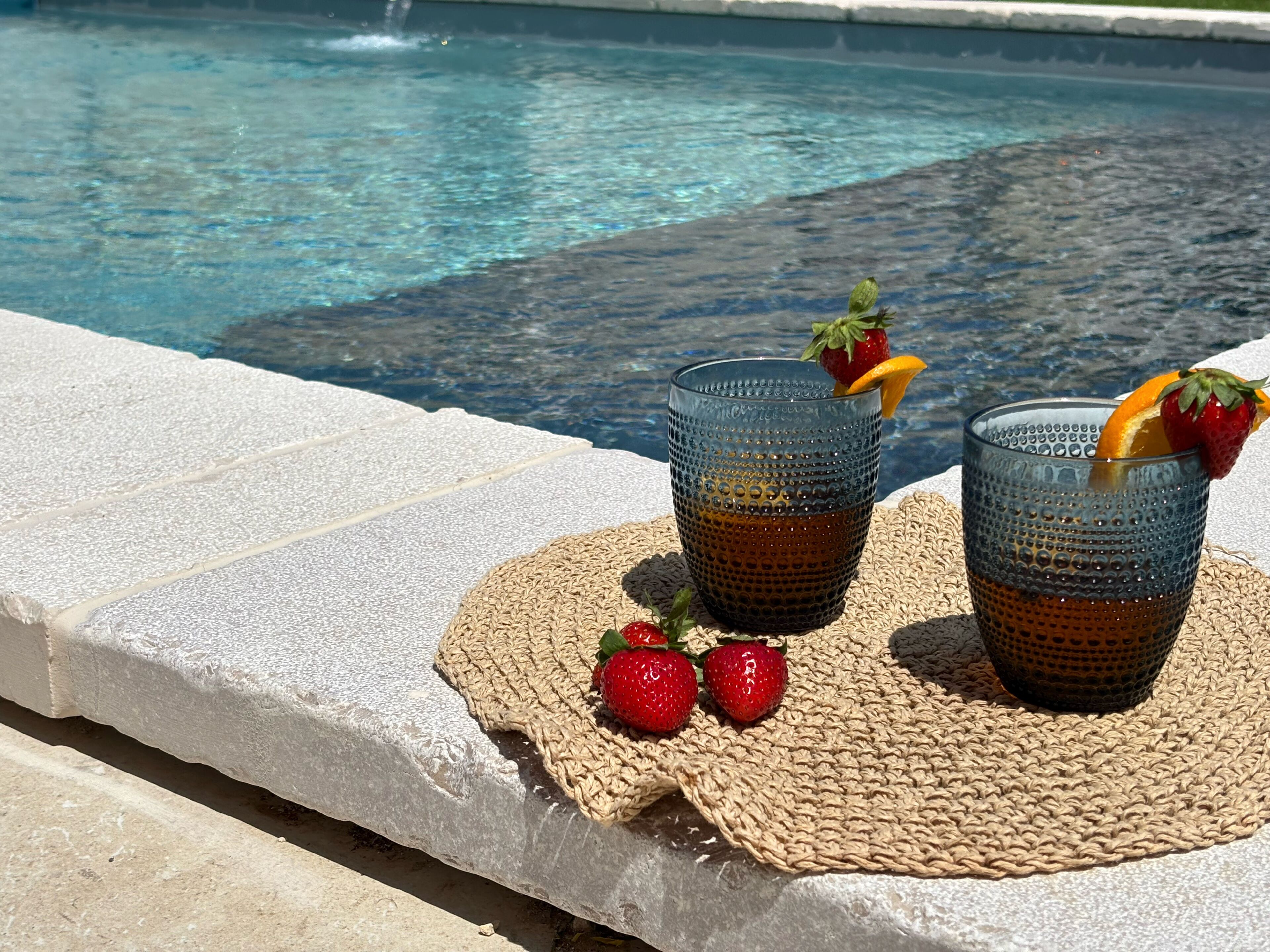Two glasses of a beverage with strawberries and an orange slice on a woven mat by a pool.