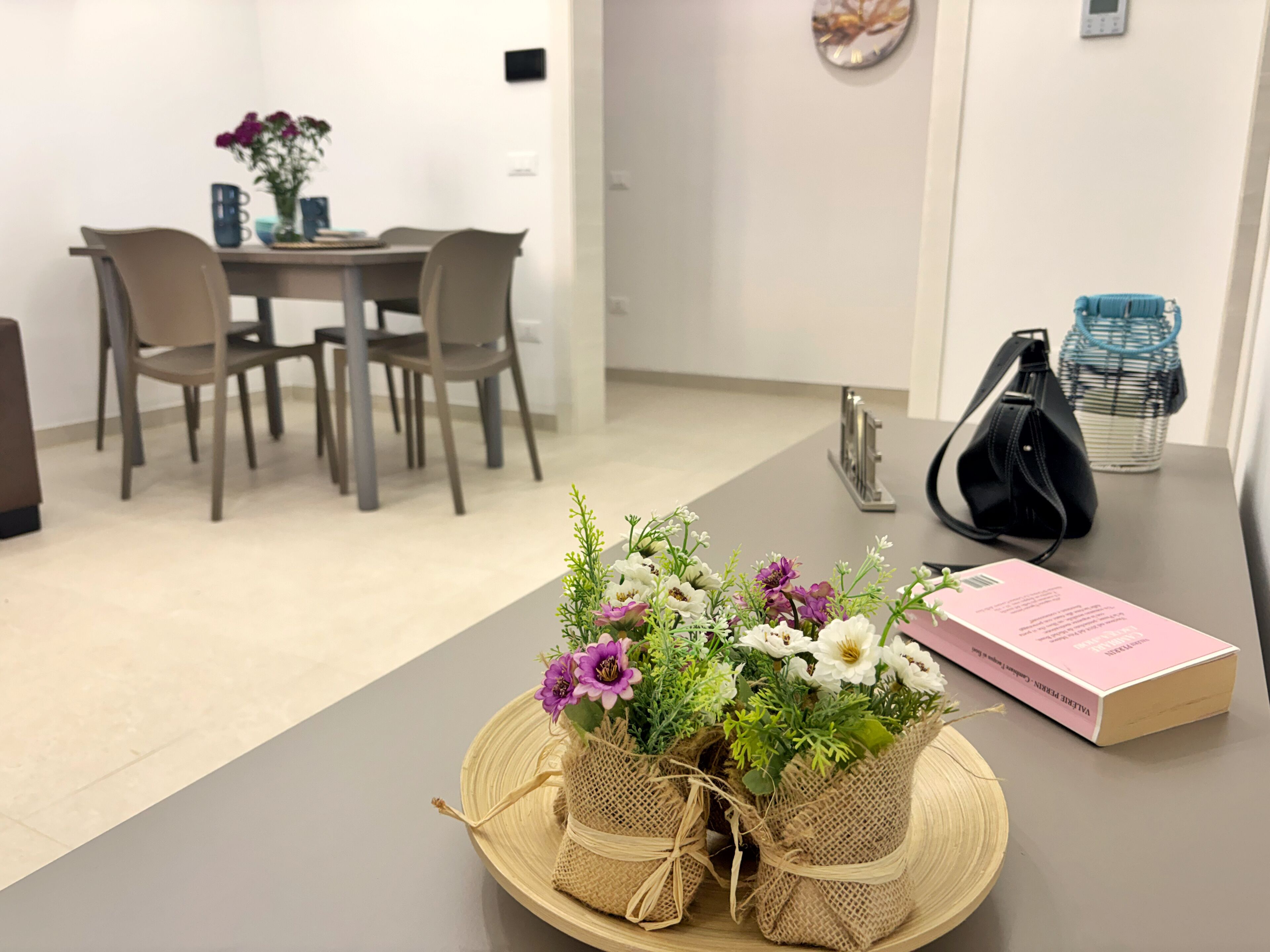 A dining area with a table set for two, a vase with flowers, a black handbag, and a pink book.