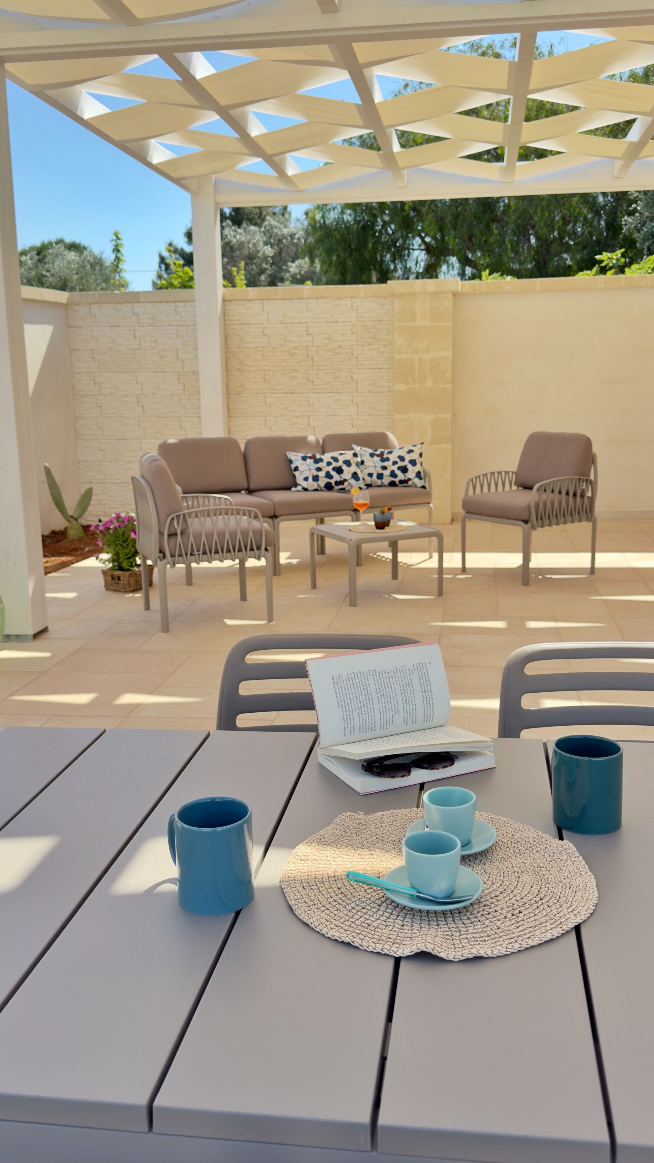 Un espacio al aire libre con una pérgola, sillas con cojines, una mesa con un libro y una bebida.