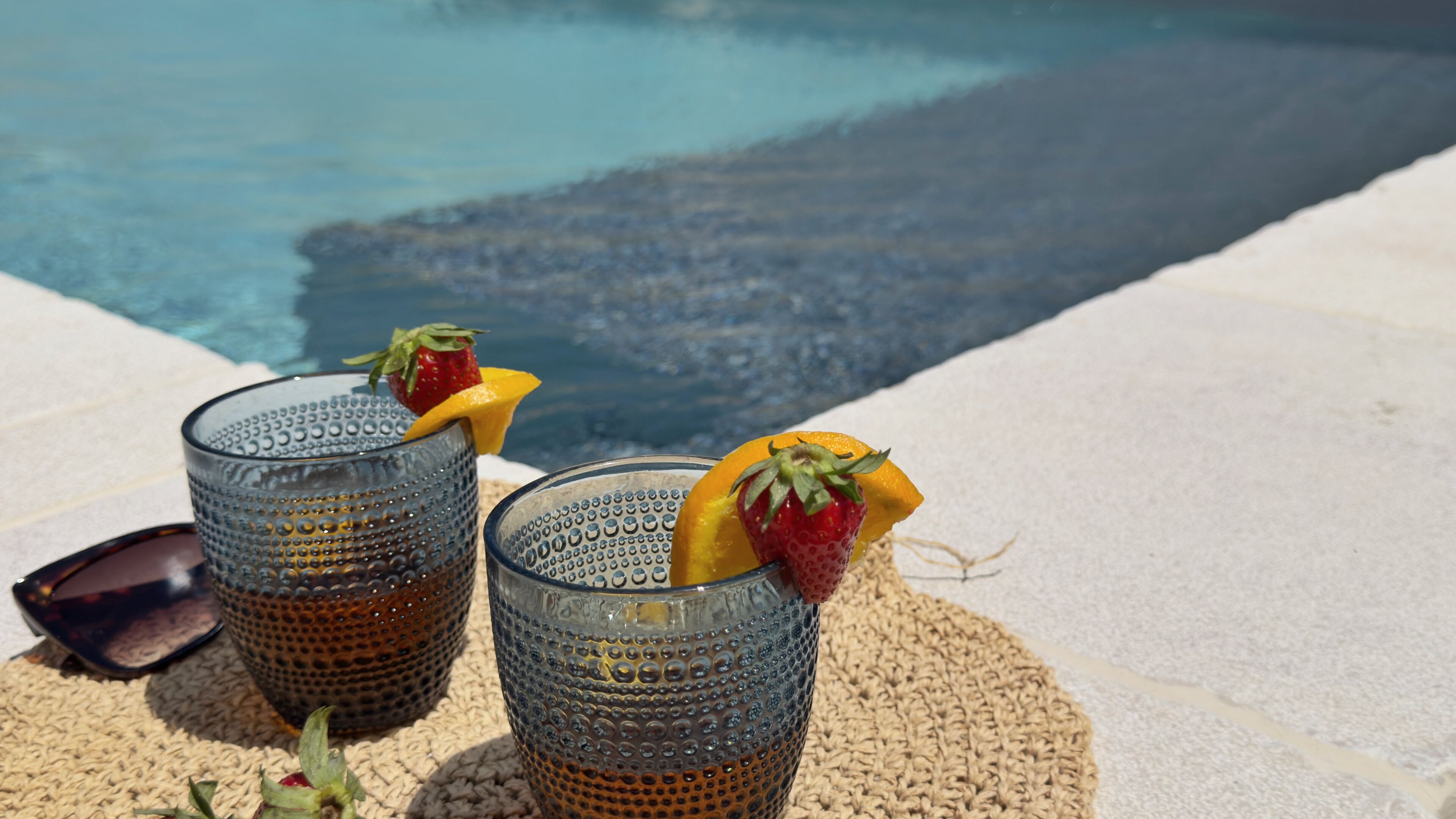 Two glasses with fruit garnishes on a textured mat near a pool.