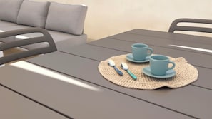 A table with two cups and spoons on a placemat, a grey sofa, and a plain wall in the background. - Alisìa (San Pietro In Bevagna)