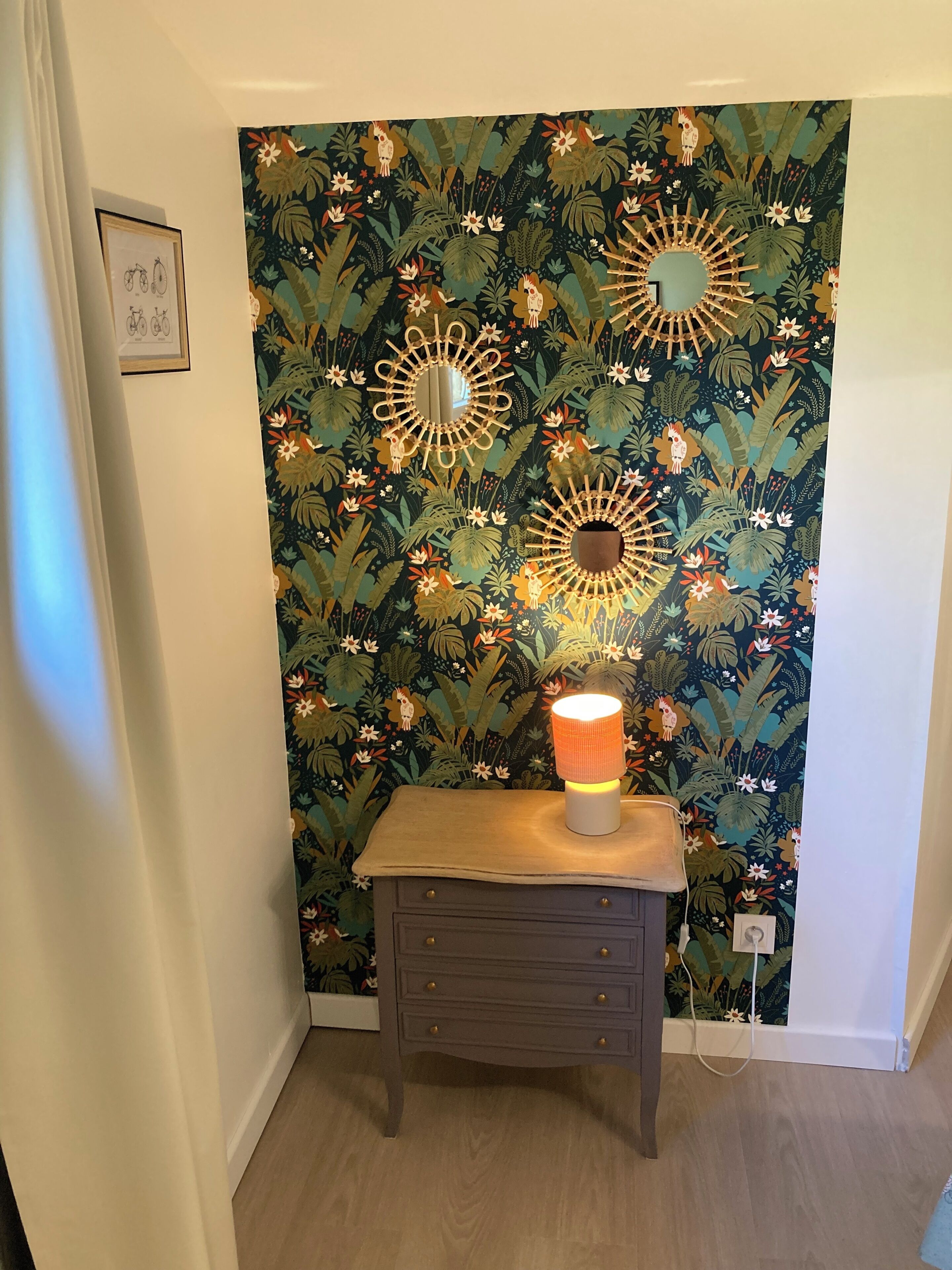 A room with a wall decorated with tropical floral wallpaper, a small wooden table with a lamp, and a framed picture on the wall.