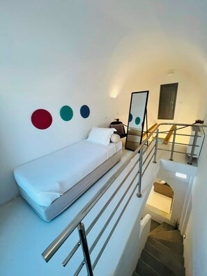 A bedroom with a single bed, a small bedside table, a chair, and a mirror. There is a staircase with a metal railing leading to an upper level. - Lagadi Suite A (Mesaria)