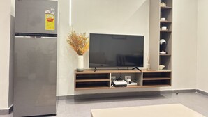 A modern living room with a flat-screen TV, a built-in wooden entertainment unit, a refrigerator, and a vase with dried flowers. - Stunning 1-Bdr Apart With Pool, Gym, Restaurant (Kinshasa)