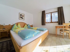 Ein Schlafzimmer mit Bett, Schreibtisch, Stuhl, Nachttisch, Lampe, einem Fenster mit Vorhängen und einem Wandbild.