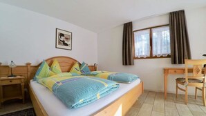 A bedroom with a bed, a desk, a chair, a nightstand, a lamp, a window with curtains, and a wall-mounted picture. - Adora Leukerbad # 4-bed apartment, Bad, WC by Interhome (Leukerbad)