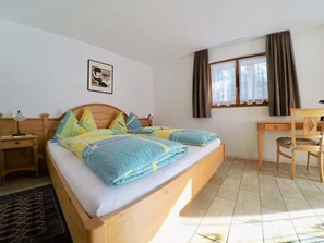 Ein Schlafzimmer mit Bett, Schreibtisch, Stuhl, Nachttisch, Lampe, einem Fenster mit Vorhängen und einem Wandbild.