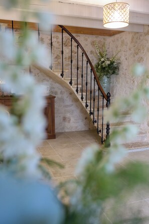 Eine geschwungene Treppe mit hölzernem Geländer und schwarzen Spindeln, die zu einem Raum mit Steinwand und einer hängenden Leuchte führt.