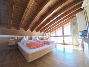 A bedroom with a sloped ceiling, wooden beams, a bed with pillows and a blanket, a bedside table, and a window with curtains. - Adora Leukerbad # 6-bed apartment, Bad, WC by Interhome (Leukerbad)