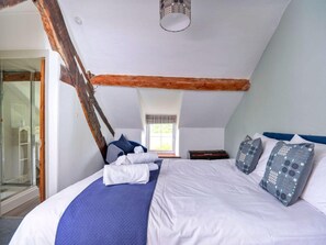 Ein Schlafzimmer mit schrägen Wänden, einem Bett mit weißer und blauer Bettwäsche, einem Fenster mit Ausblick und Holzbalken.