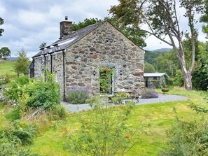 Ein Haus aus Stein mit einem geneigten Dach, umgeben von einer Rasenfläche mit verschiedenen Pflanzen und einem Kiesweg, der zum Eingang führt.