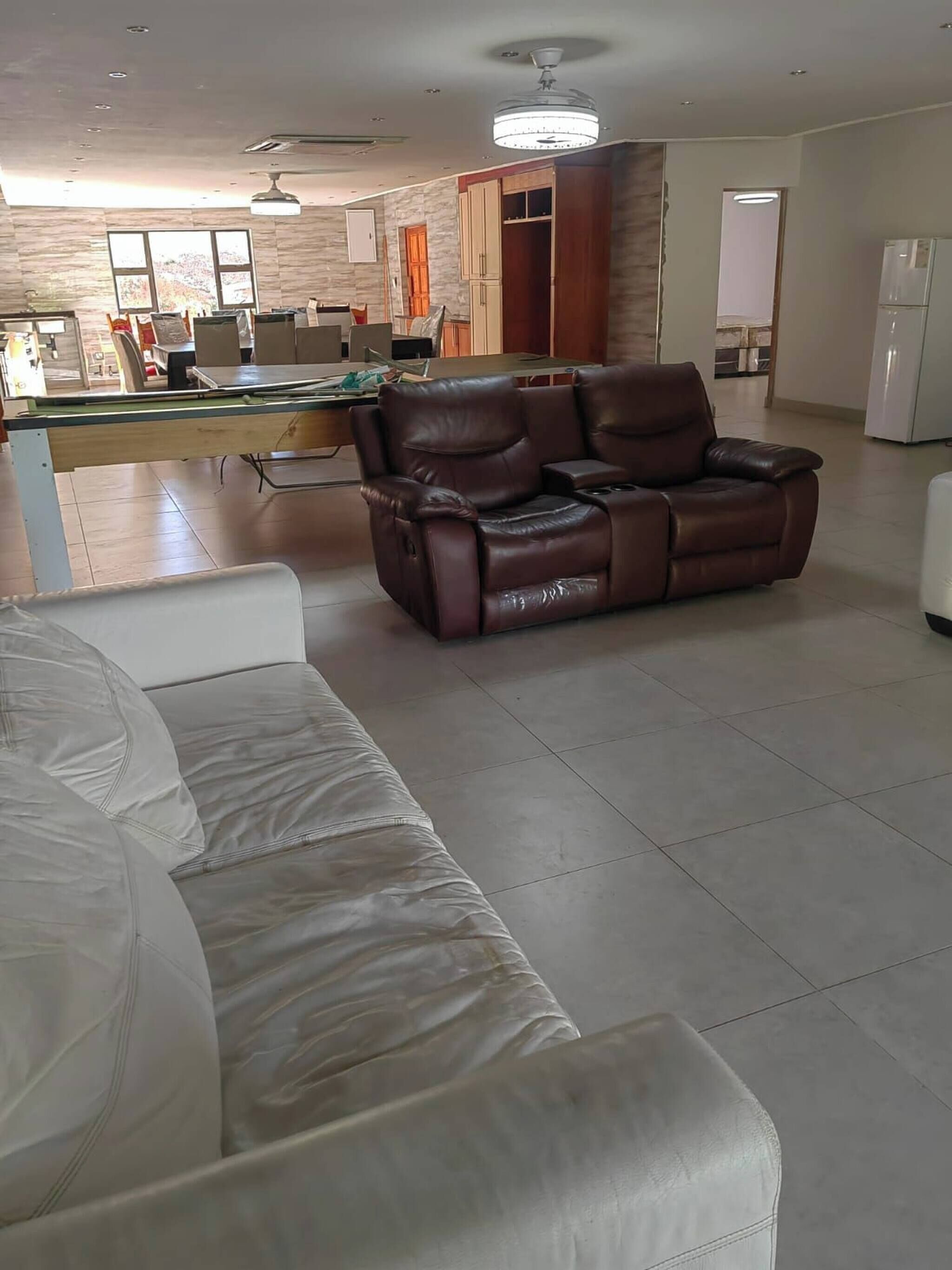 A living room with a leather sofa, a white sofa, a dining area with a table and chairs, and a kitchen in the background.
