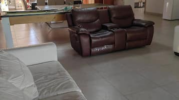 A living room with a leather sofa, a white sofa, a dining area with a table and chairs, and a kitchen in the background.