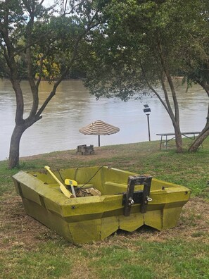 A green boat with a paddle and a chair on a grassy area near a body of water, with trees and a picnic table in the background. - Luxurious Riverside Retreat in Lindequesdrif, Vaal River (Lindequesdrif)