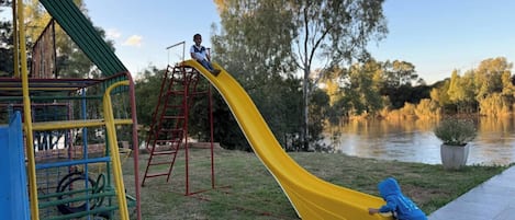 Un parque infantil con un tobogán amarillo, una estructura para escalar y un cuerpo de agua al fondo.