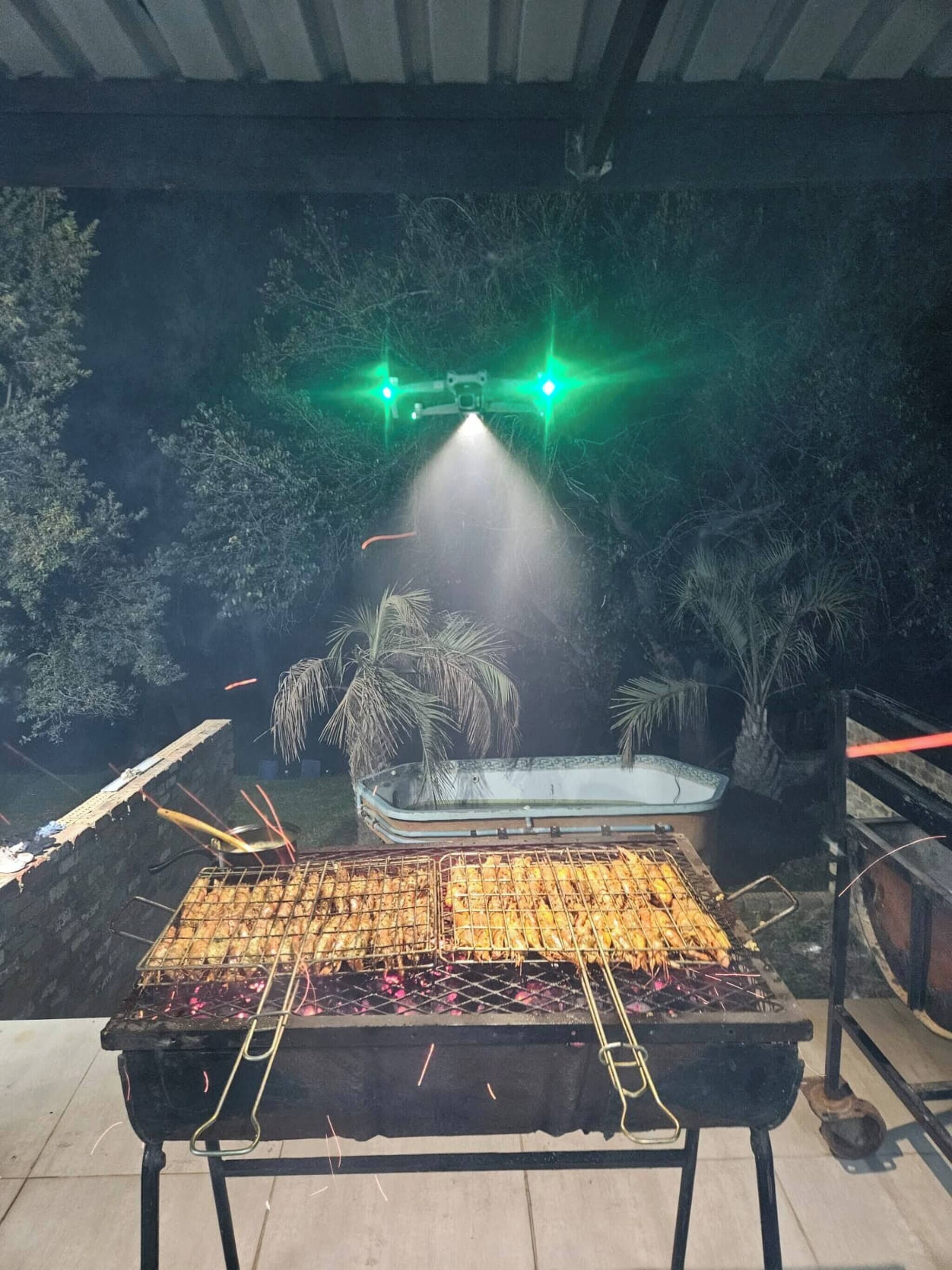 A barbecue grill with skewers of food, a hot tub, and a green-lit drone overhead.