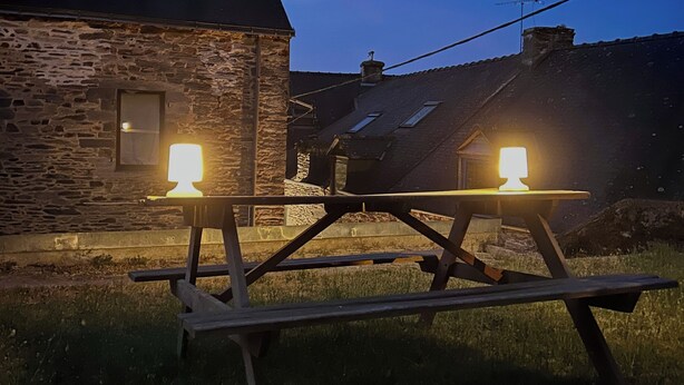 A stone building with a pitched roof, a wooden picnic table with two lights, and a grassy area. - Colorful 3-bedroom apartment in the heart of downtown with outdoor space (La Gacilly)