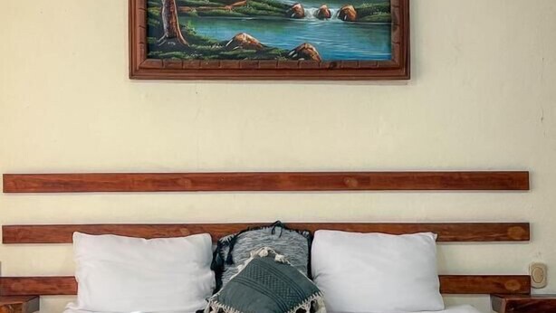 Una cama con sábanas blancas, cabecera de madera y un cuadro de un paisaje tropical.