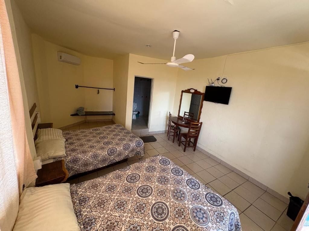 Ein Zimmer mit zwei Betten, einem Deckenventilator, einem an der Wand befestigten Fernseher und einem kleinen Tisch mit Stühlen.