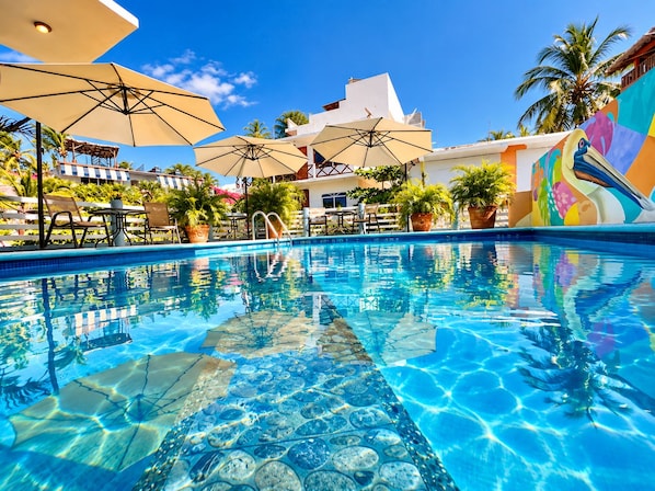 A swimming pool with clear water, surrounded by lounge chairs and umbrellas, and a colorful mural on the adjacent wall. - Casa piedra parada hotel (Brisas de Zicatela)