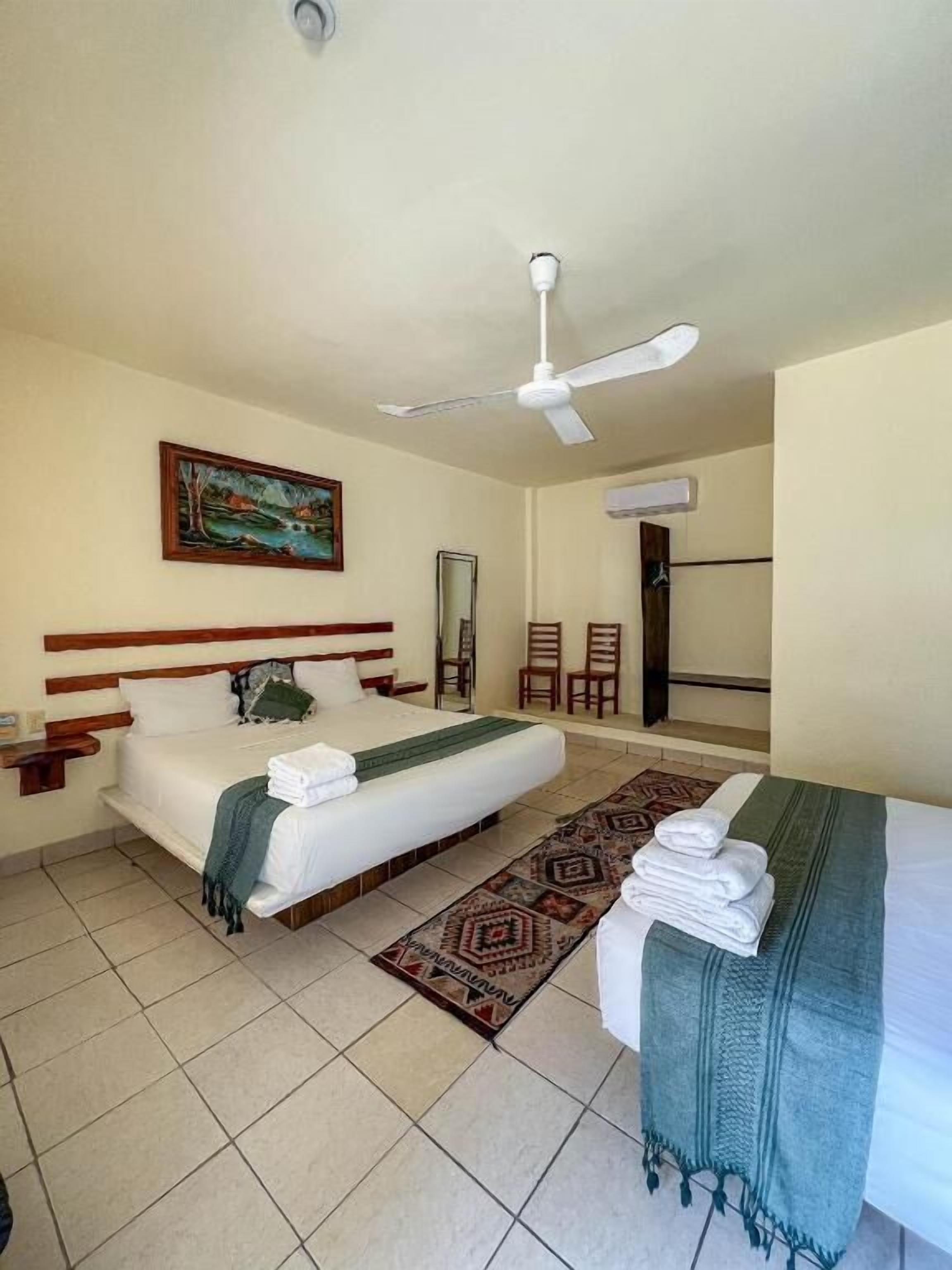 Habitació d'hotel amb llit, ventilador de sostre, quadre a la paret i taula petita amb cadires.