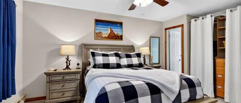 A bedroom with a bed, a nightstand, a lamp, a ceiling fan, a painting, and a closet.